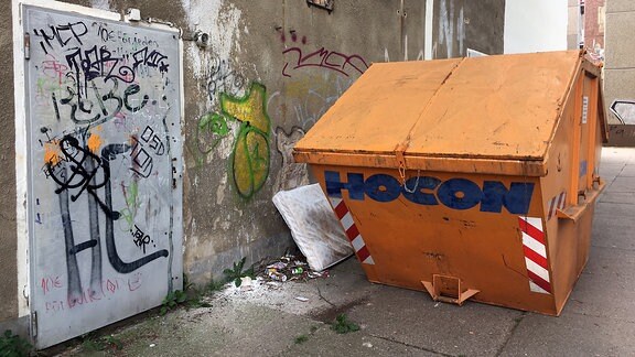 Eine Häuserwand ist mit Graffitis beschmiert, eine alte Matratze liegt hinter einem Behälter für Bauschutt