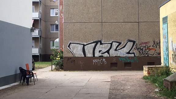 Eine Häuserwand ist mit Graffitis beschmiert, drei Plastikstühle stehen vor alten Wohnblocks