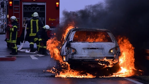 Ein Auto steht in Flammen. Im Hintergrund  befinden sich drei Feuerwehrmänner an einem Löschfahrzeug