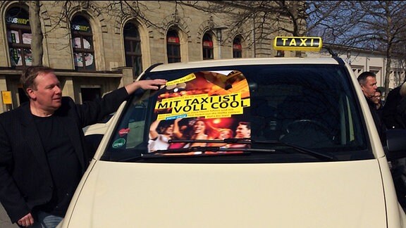 Auf der Frontscheibe eines Taxis liegt ein Plakat mit der Aufschrift: Mit Taxi ist voll cool. Die Aktion "Fifty-Fifty-Taxi" wird trotz gestiegener Preise fortgesetzt