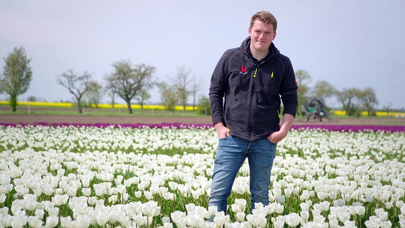 Ein junger blonder Mann in Jeans und Pullover steht inmitten weißer Tulpen auf einem Feld 