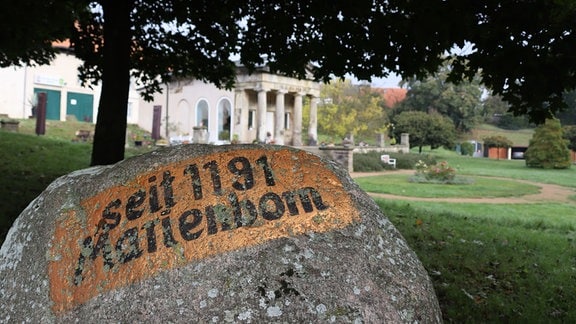 Auf einem großen Grauen Stein steht "seit 1191 Marienborn", im Hintergrund ist die Orangerie von Marienborn