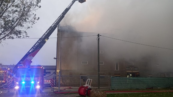 Aus dem Dachstuhl eines Gebäudes in Osterweddingen dringt dichter Rauch, eine Drehleiter der Feuerwehr ist zu sehen.