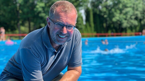 Ein Mann hockt in einem flachen Becken eines Freibades und lächelt in die Kamera, im Hintergrund planschen Kinder.