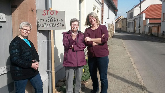 Drei Frauen stehen an einer Straße. An einem Haus ist ein Schild aufgestellt, dass den Stopp der Straßenausbaubeiträge fordert.