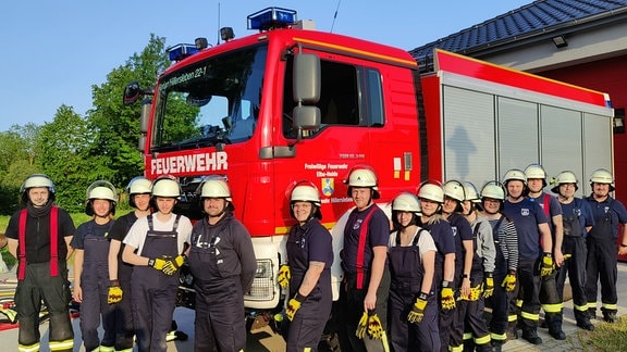 Männer und Frauen in Feuerwehruniform stehen vor einem rot glänzenden Löschfahrzeug