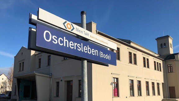 Vor einem Bahnhof steht ein blaues Schild mit der Aufschrift 'Oschersleben (Bode)'.