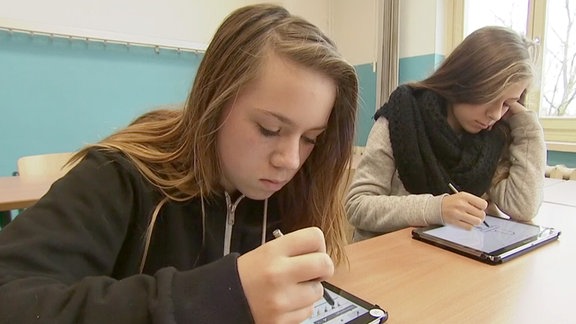  Zwei Mädchen sitzen mit Tablet im Klassenzimmer.