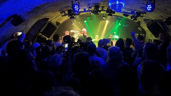 Blick in Fischaugenoptik durch einen dunklen Clubraum über die Schatten des Publikums hinweg nach vorne auf die beleuchtete Bühne, auf der eine Rockband spielt