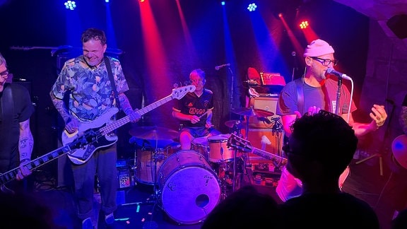 Eine kleine violett beleuchtete Konzertbühne mit vier Musikern: einem Sänger am Mikrofon, einem Gitarristen, einem Bassisten und einem Schlagzeuger.