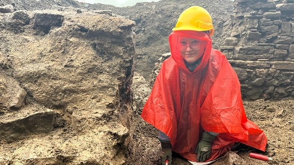 Eine junge Frau mit Helm und Regencape kniet in einer Ausgrabungsstelle.