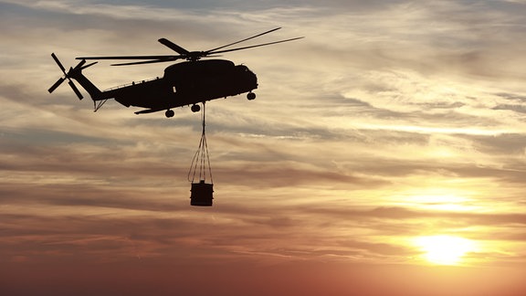 Ein Hubschrauber der Bundeswehr fliegt im Licht der untergehenden Sonne mit einem Wasserbehälter zum Königsberg am Brocken. 