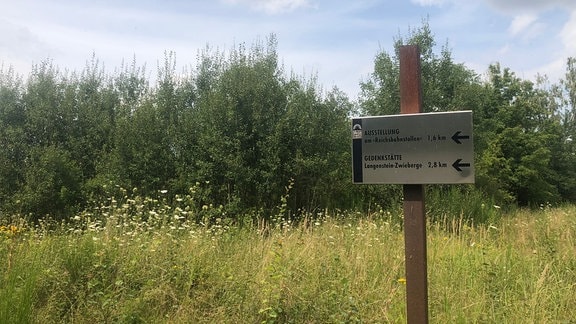 Ein Wegweiser auf einer Wiese zeigt mit Pfeilen in Richtung der KZ-Gedenkstätten-Ausstellung