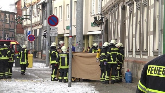 Mehrere Feuerwehrleute verdecken mit einer Plane den Eingang eines Fachwerkhauses in Halberstadt, in der es eine Explosion gab.