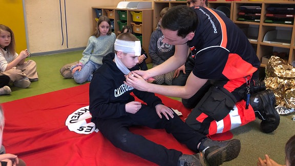 Kinder sitzen im Kreis auf dem Boden. Im Kreis zeigt ein Mitarbeiter des Roten Kreuzes an einem Kind, wie man einen Verband anlegt.