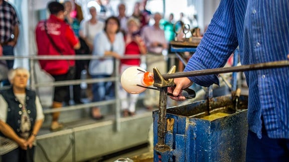 Ein Glashandwerker in blauer Arbeitskleidung bearbeitet ein glühendes Stück Glas an einem langen Stab.