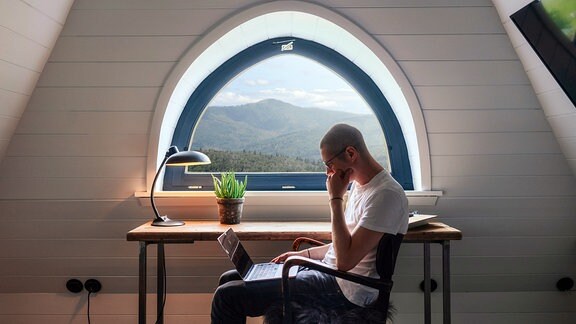 Ein Mann sitzt mit einem Laptop auf dem Schoss in einem Zimmer mit Aussicht über einen Wald