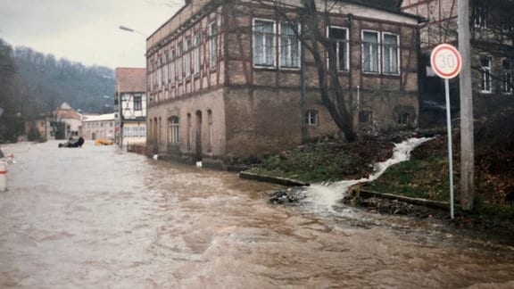 Häuser stehen direkt an einer überfluteten Straße, daneben fließt ein kleiner Bach die Wiese hinunter. Das Bild sieht analog aus.