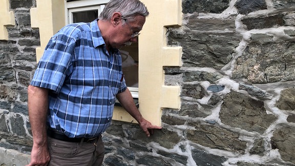 Ein älterer Mann steht vor einem Haus und hält die Hand an das untere Ende eines Fensters
