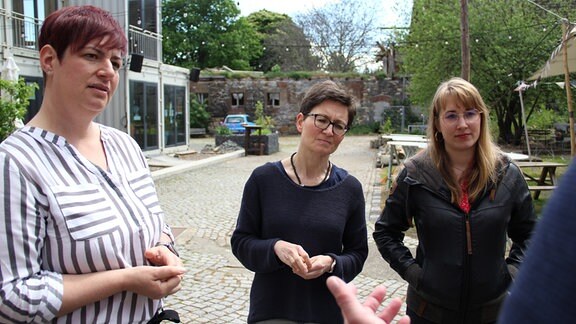 Drei Frauen stehen in einer Runde auf einem Hof und unterhalten sich. 