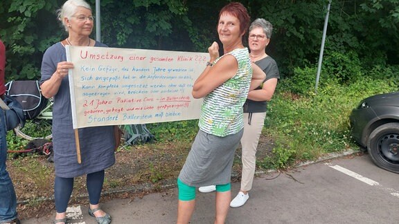 Drei Frauen halten ein Schild mit einer Protest-Botschaft in die Kamera.