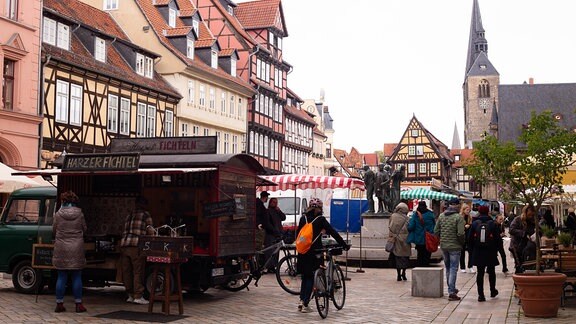 Ein Platz, auf dem Verkaufsbuden stehen, Fachwerkhäuser im Hintergrund, ein paar Menschen sind unterwegs