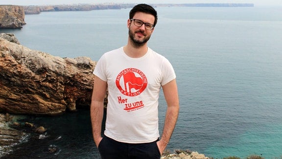 Ein Mann mit dunklen kurzen Haaren und Vollbart mit Brille steht in weißem Antifa-T-Shirt auf einem Felsen vor einer Meeresbucht