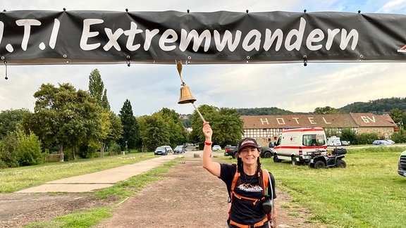 Eine Frau steht unter einem Schild mit der Aufschrift "Extremwandern" und läutet eine Glocke