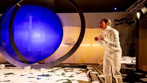 Solaris, Eine Frau im Weltraumanzug steht auf einer Bühne, vor ihr liegen viele Zettel auf dem Boden, im Hintergrund ein großer Kreis, der blau beleuchtet ist.