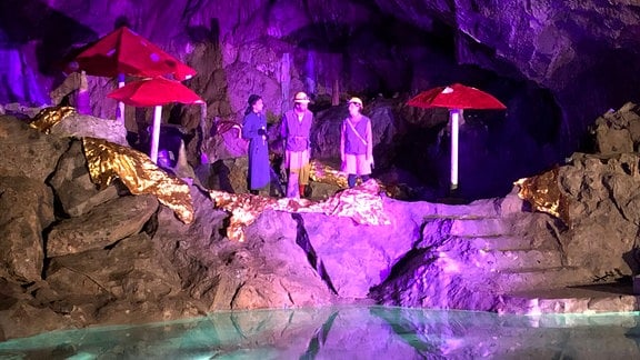 Drei Schauspieler stehen in einer Höhle, vor ihnen ist Wasser, sie werden von pinkem Licht angestrahlt.