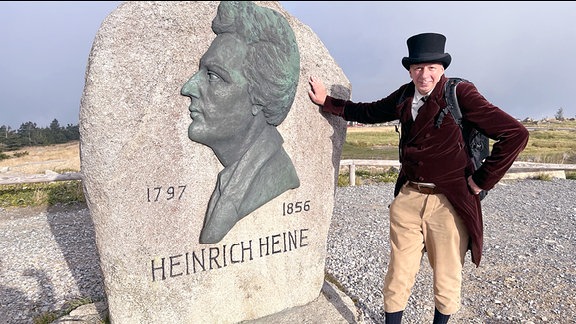 Der Wanderer Michael Lütje als Heinrich Heine steht an einem Heinrich-Heine-Gedenkstein.