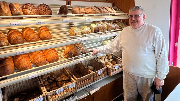 Bäcker steht vor Regal mit Broten und Brötchen und lächelt