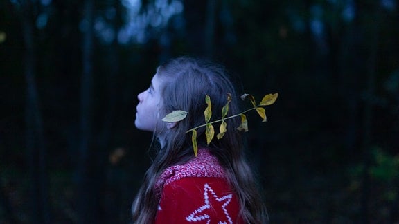 Ein Kind mit roter Jacke und langen Haaren hat einen Zweig im Haar und steht im Wald