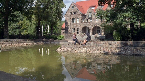Zwei Menschen sitzen vor der Kulisse einer altehrwürdigen Villa