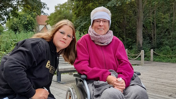 Eine junge Frau mit Brille sitzt neben einer älteren Frau im Rollstuhl. 
