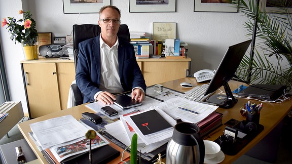 Andreas Henke, ein Mann im Anzug mit schütterem Haar und Brille, sitzt an einem Schreibtisch. Darauf liegen viele gestapelte Papiere und ein Handy. Am Rand stehen eine Kaffeetasse und eine Kanne.