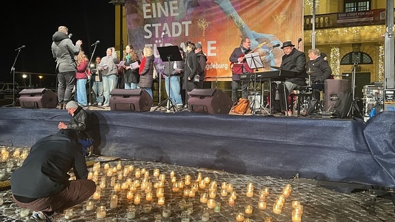 Menschen singen auf einer Bühne auf dem alten Markt von Magdeburg Friedenslieder anlässlich der Zerstörung Magdeburgs am 16. Januar 1945. Davor zünden Menschen Kerzen an.