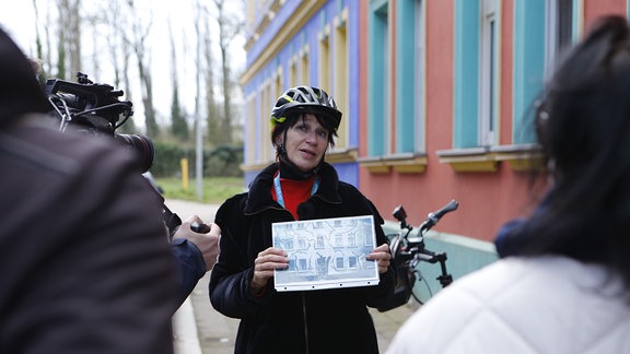Eine Frau mit einem Fahrradhelm steht vor einem bunten Haus und zeigt anderen Menschen ein Bild, das sie in ihrer Hand hält.