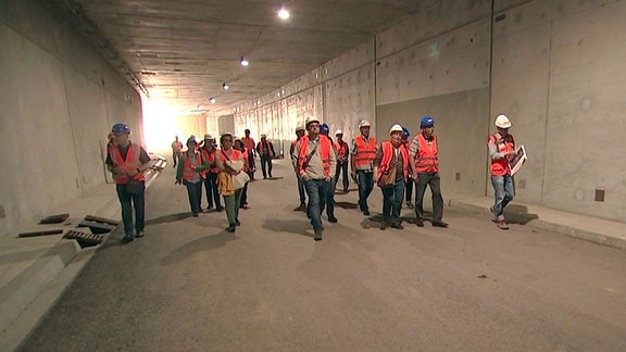 Menschen mit Bauhelmen und Warnwesten laufen durch einen Tunnel. 