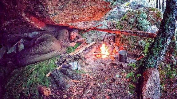 Ein Mann liegt in einer Felsspalte, neben sich ein Feuer