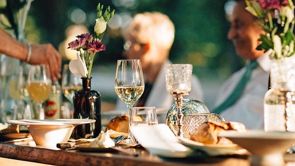 Ein festlich gedeckter Tisch mit Weingläsern, im Hintergrund verschwommen eine blonde Frau und ein Mann