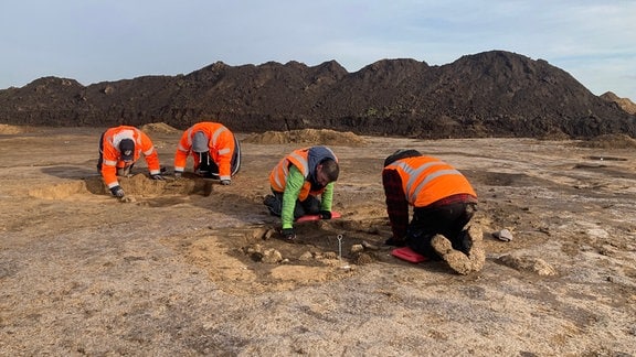 Mehrere Archäologen in orangenen Warnwesten knien bei Grabungen vor dem Bau der Intel-Chip-Fabrik auf dem Boden.