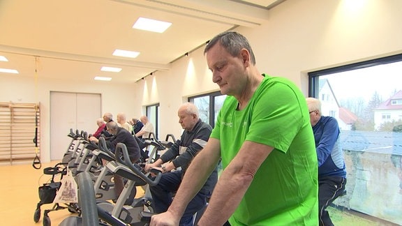 Mann in grünem T-Shirt trainiert auf einem Fitnessfahrrad in einem Raum mit mehreren alten Männern zusammen.
