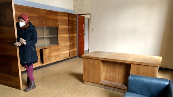 Eine der Projektleiterinnen zeigt das Chefarzt-Zimmer mit alten Sofas, Tisch und Schrankwand.