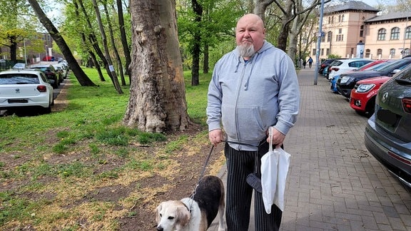 Ein Mann mit einem grauen Kapuzenpullover und einem Hund an der Leine steht neben Autos auf dem Bürgersteig