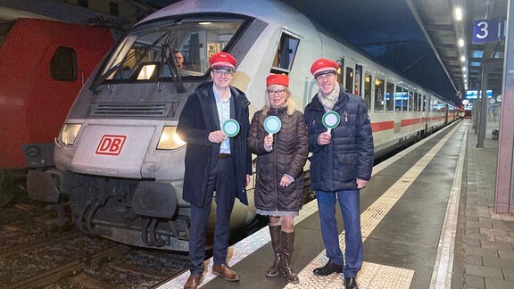 zwei Männer und eine Frau mit Bahnmützen und einer grünen Kelle stehen vor einem weißen Zug. 