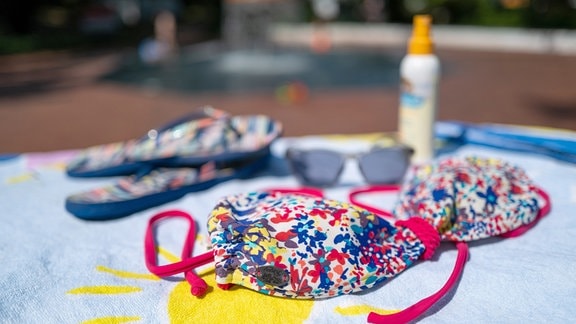 Ein Bikini-Oberteil liegt neben einer Sonnenbrille, Flip Flops und Sonnencreme auf einem Badehandtuch