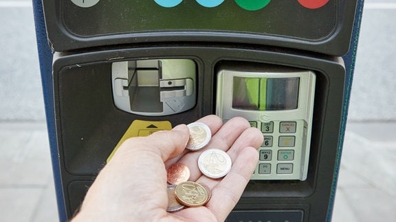 Eine Hand hält Euromünzen vor einem Parkscheinautomaten.
