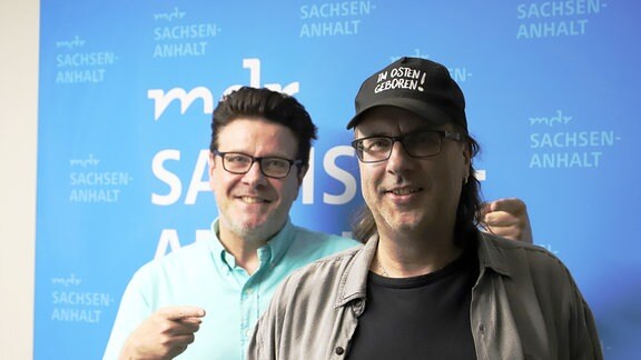 Dunkelhaariger Mann mit Brille in hellblauem Hemd und langhaariger Mann mit schwarzem Käppie vor einem MDR SACHSEN-ANHALT-Logo