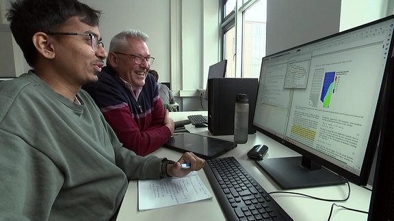 Ein lachender Mann sitzt neben einem Studenten an einem Computer.